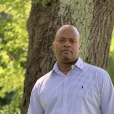 A person with a shaved head is standing in front of a tree, looking into the camera.
