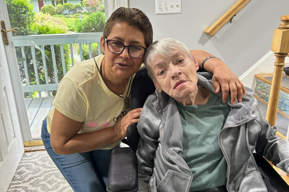 A middle aged adult with short hair and glasses embraces a senior adult using a wheelchair. They are posing for a picture in a home's hallway.