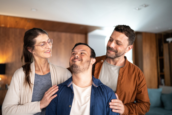 Two middle-aged parents look fondly at their adult child with an intellectual disability. All are smiling.