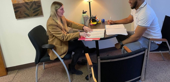 A light skinned person with long blond hair and dark skinned person with short dark hair work together at a table.