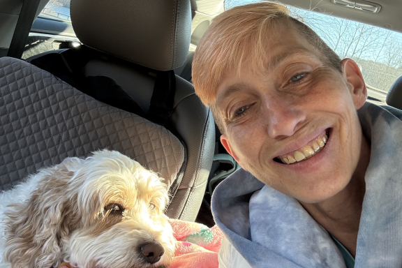 An adult with short blond hair takes a smiling selfie in the car with her fluffy dog