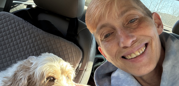 An adult with short blond hair takes a smiling selfie in the car with her fluffy dog