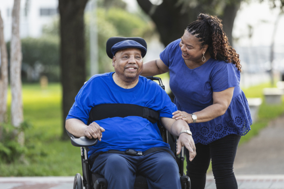 Two adults outdoors smiling. One is using a wheelchair, and the other is leaning into him.