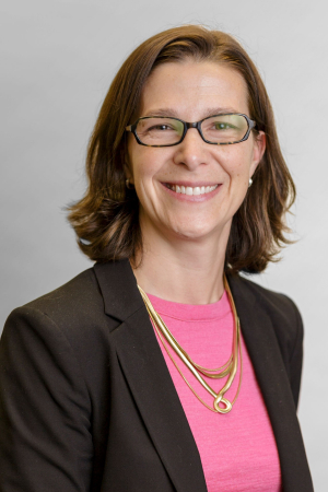 A light-skinned adult with shoulder length brown hair and glasses wears a pink shirt and dark blazer and smiles at the camera in a formal headshot.