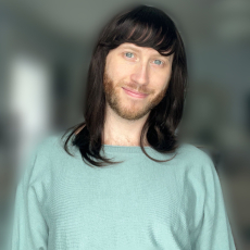 A person with shiny long dark hair, light skin and eyes, and a beard smiles at the camera.