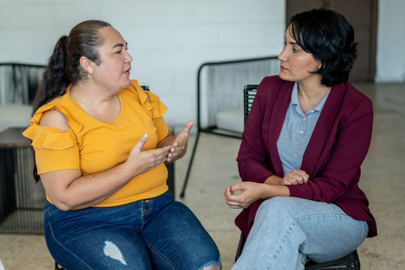 Two adults sitting in chairs have a conversation. One is dressed casually, and the other is in business casual.