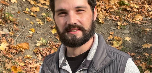 A man with light skin and dark hair smiles at the camera in a fall outdoor setting.