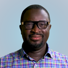 An adult with dark skin, short hair, and a collared plaid shirt smiles at the camera